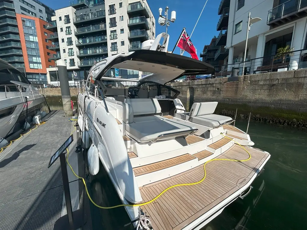 Luxury boat docked at a marina, featuring a flag, spacious seating, and modern buildings in the background under clear blue skies.